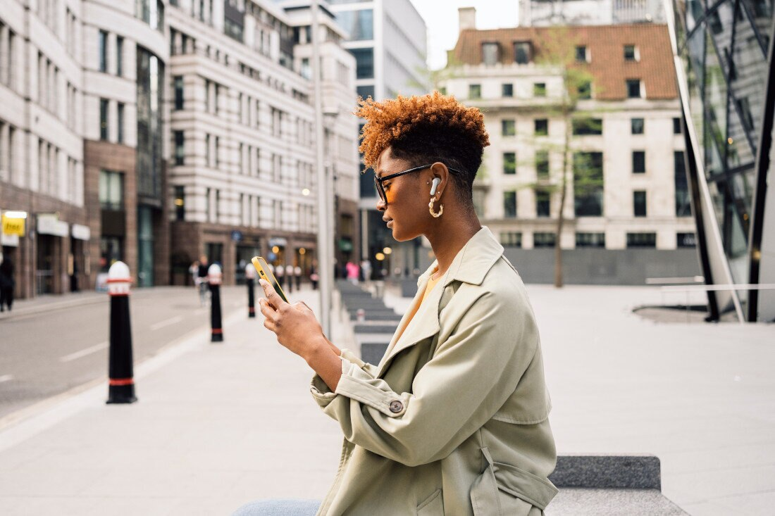 Kobieta z afro i okularami przeciwsłonecznymi, korzystająca z telefonu, stojąca w mieście. Budynki za nią.