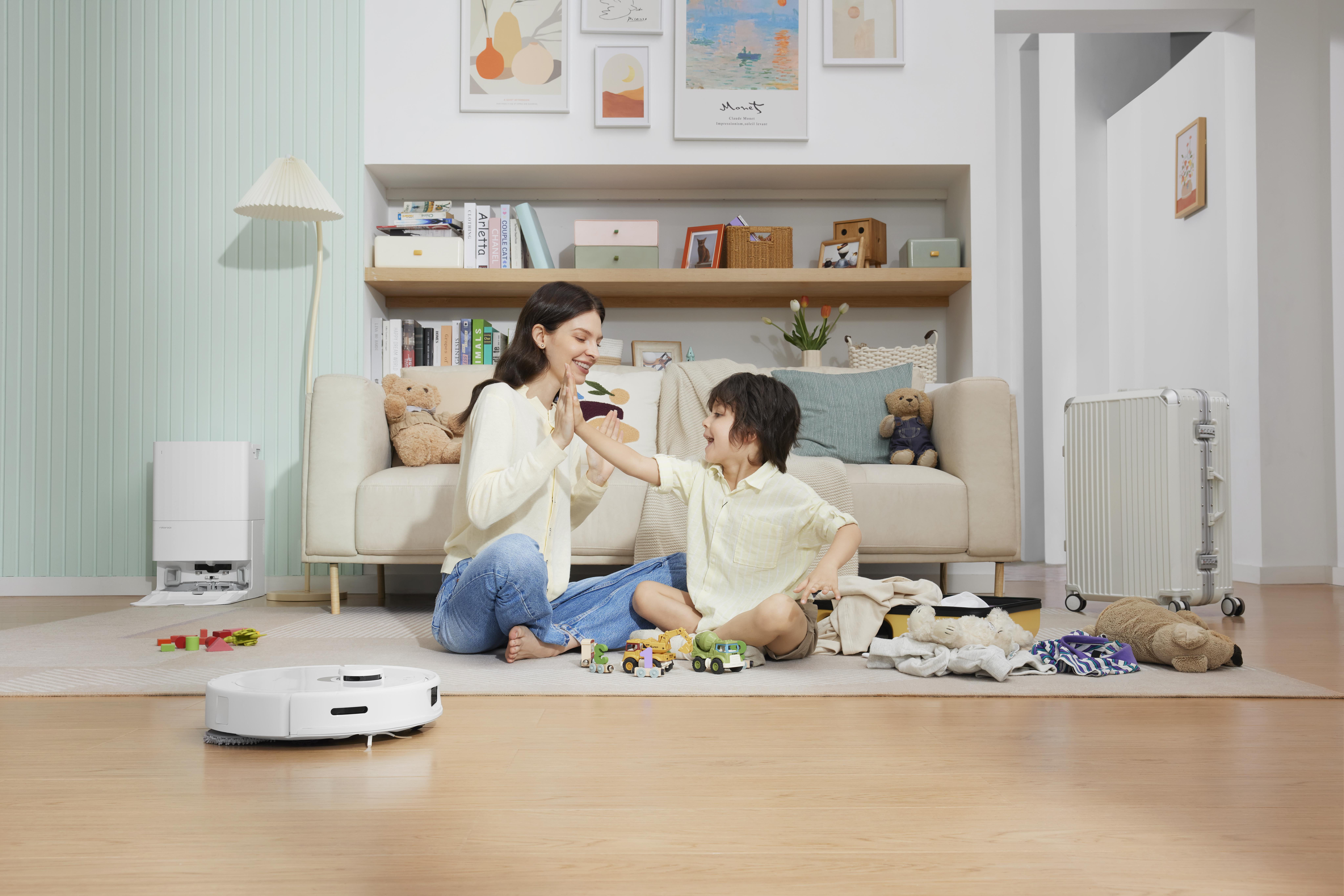 Eine Frau und ein Junge spielen mit einem Roboter-Staubsauger auf dem Boden. Sofa und Regale im Hintergrund.
