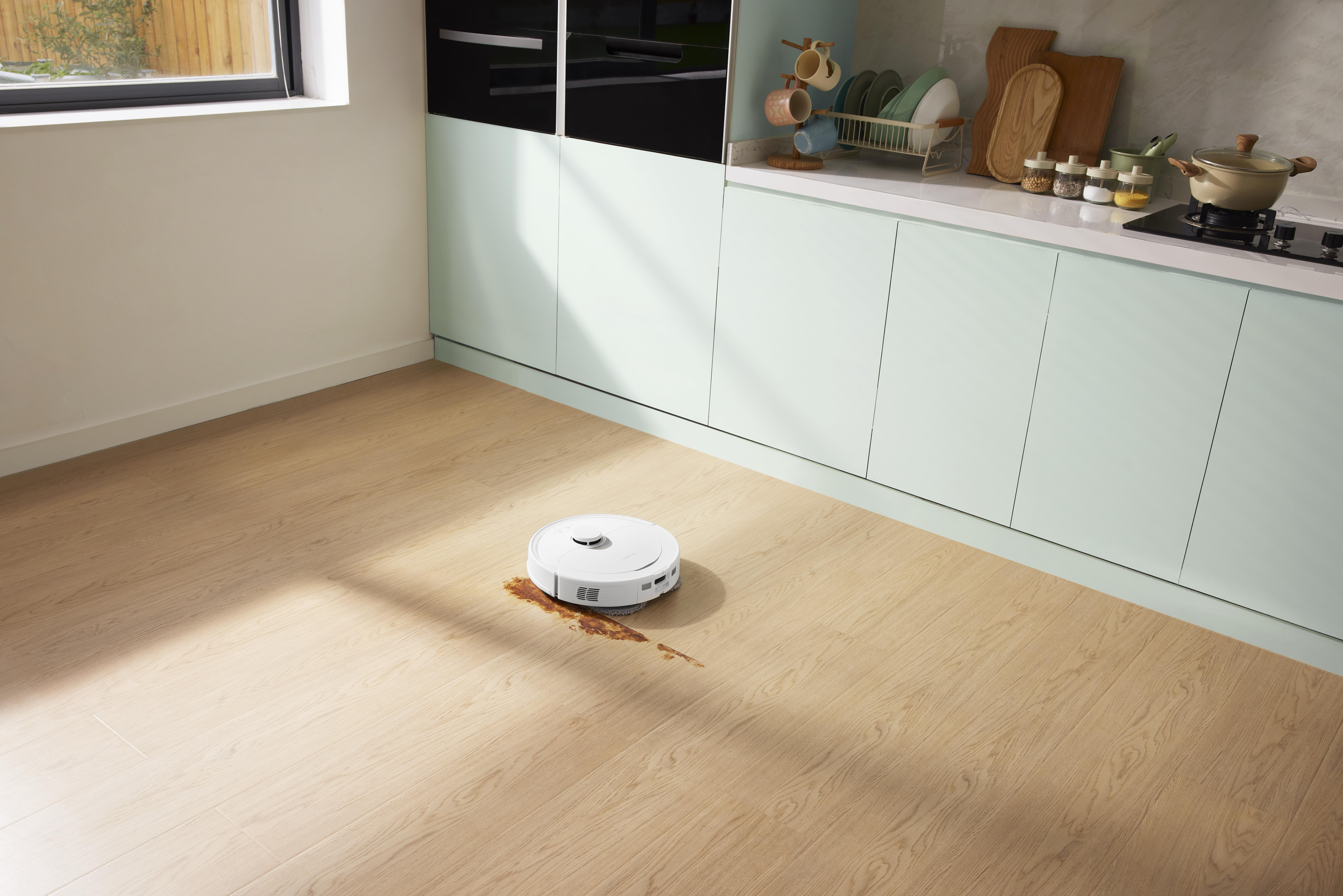 Roboter-Staubsauger auf Holzboden mit Fleck, Küchenschränke im Hintergrund.