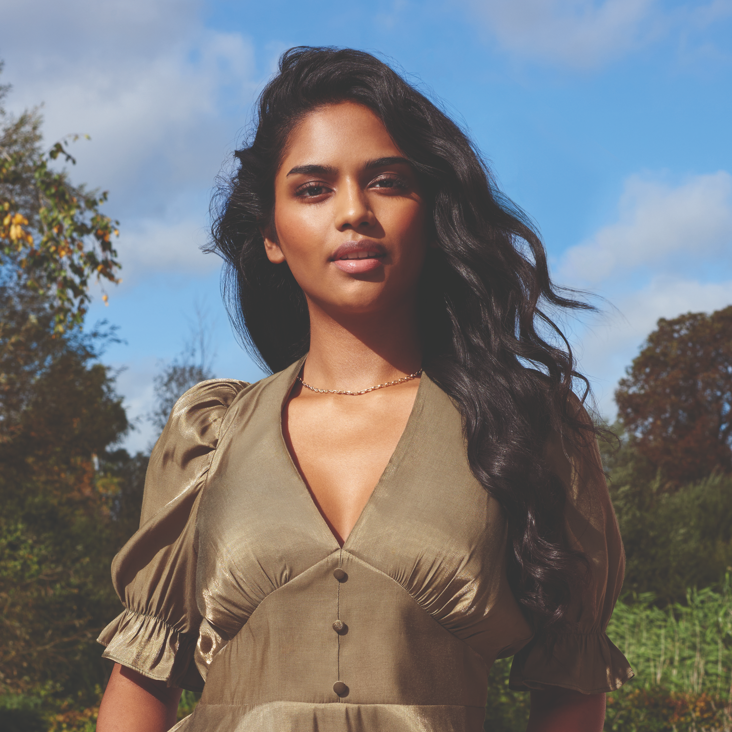 Woman in a green dress, looking up with a smile, trees and sky in the background.