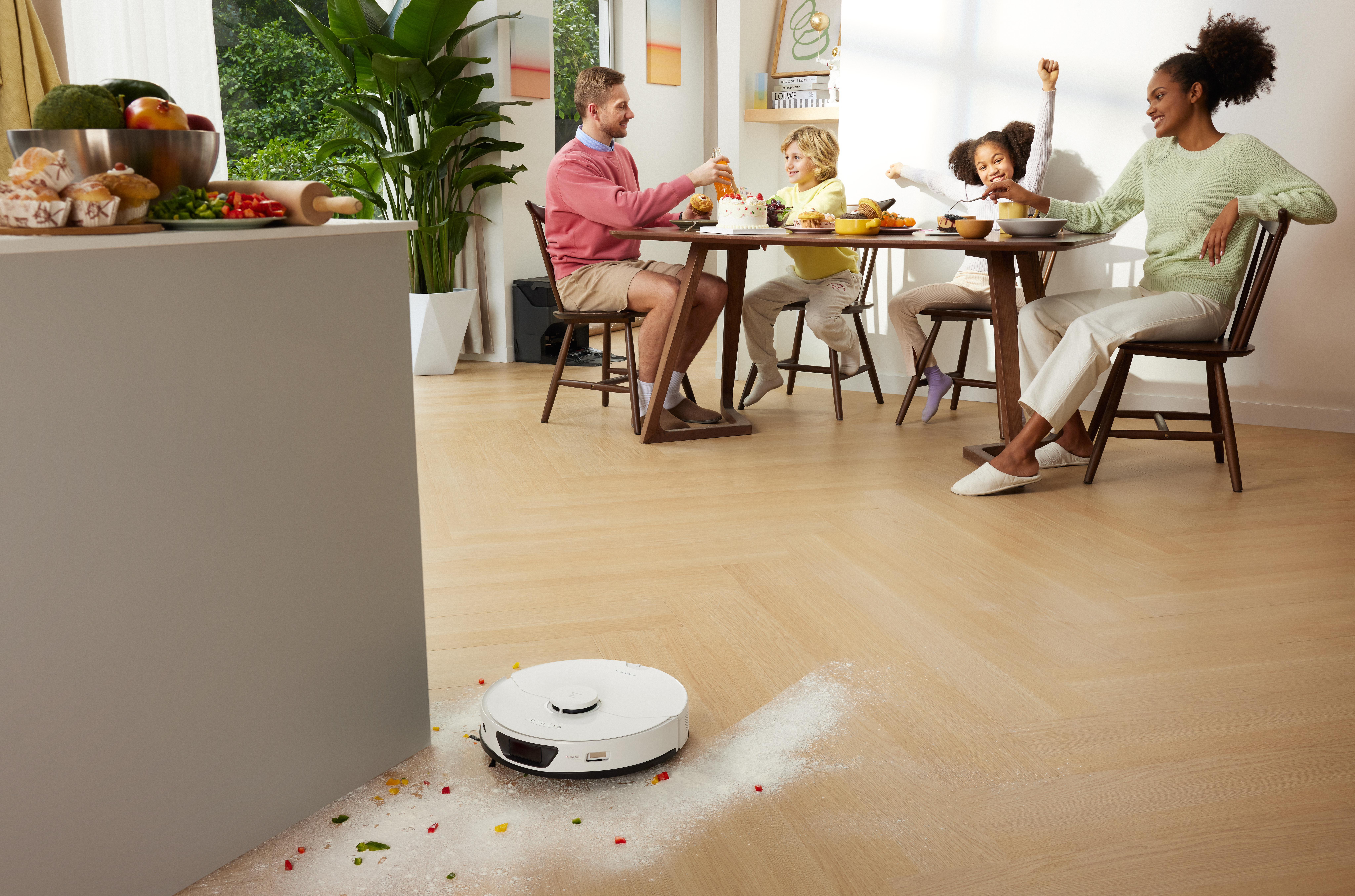 Eine Familie genießt eine Mahlzeit, während ein Roboterstaubsauger den Holzboden reinigt.