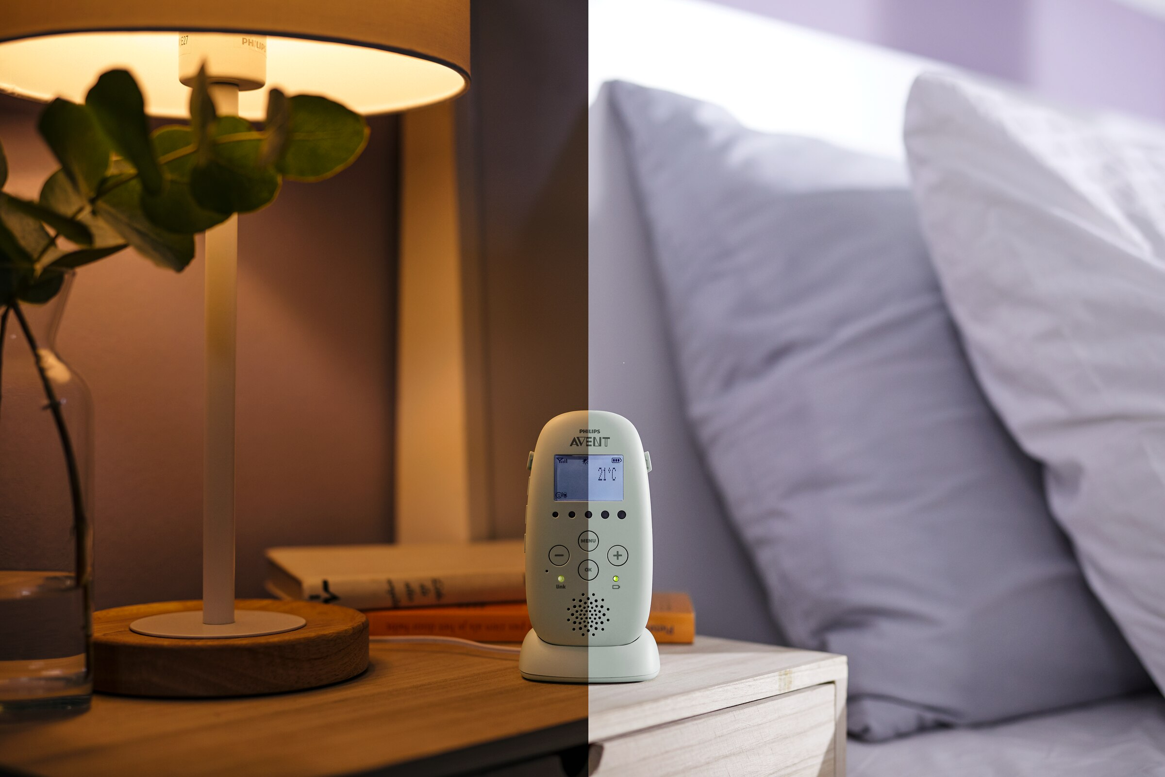 Niania elektroniczna na stoliku nocnym. Lampa po lewej, łóżko z poduszkami po prawej.