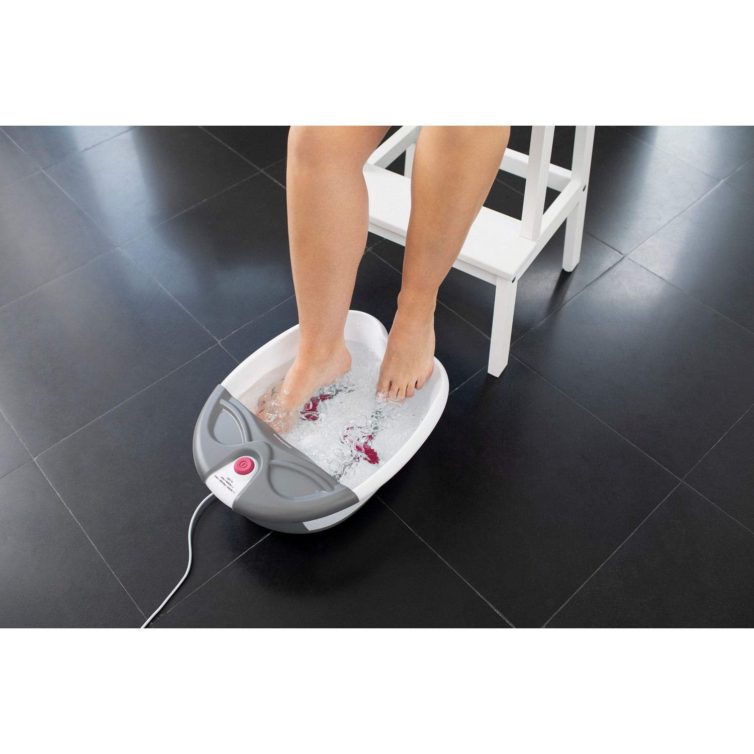 Pieds d'une personne dans un bain de pieds, eau avec des bulles. Tabouret blanc et sol carrelé noir.