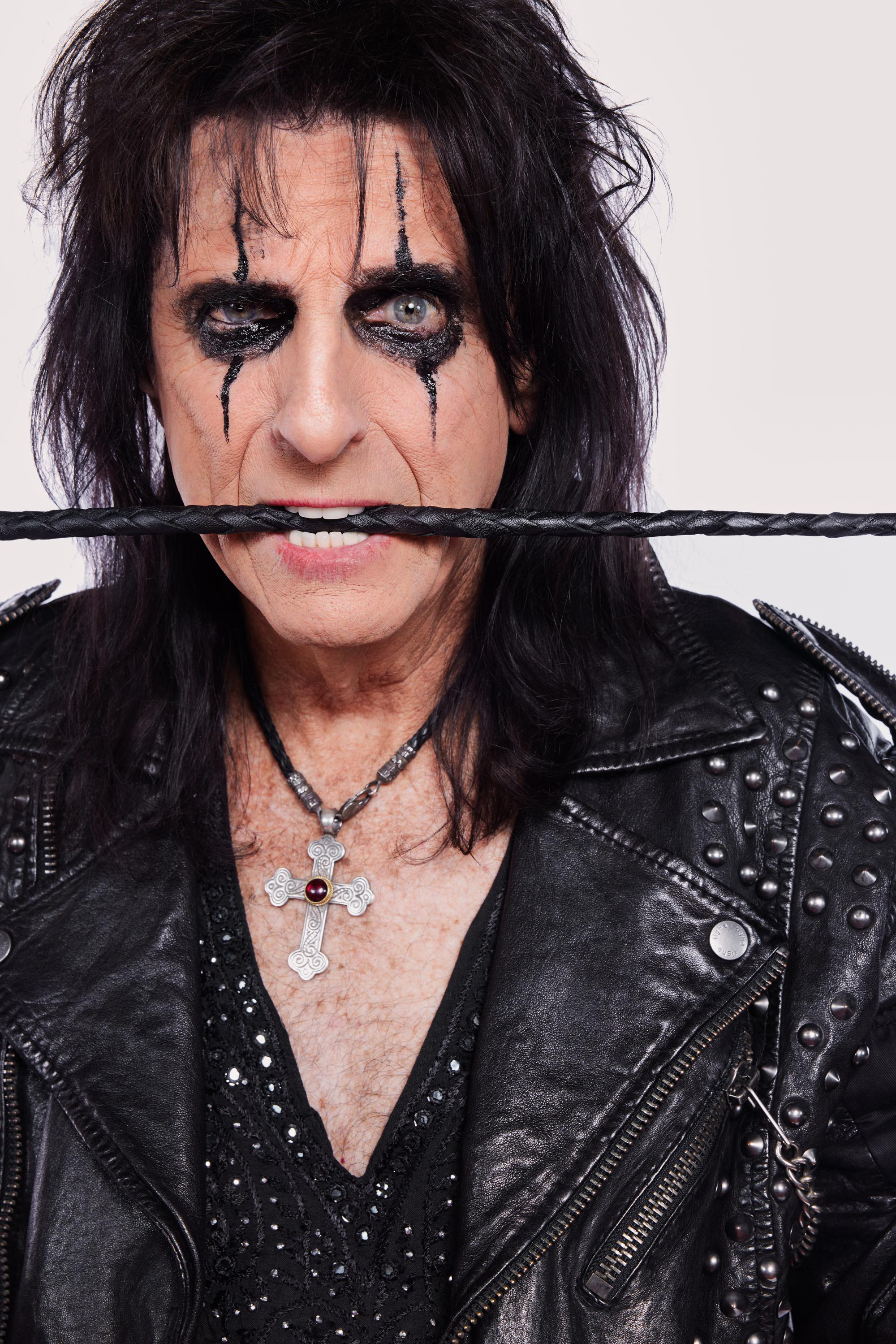 Alice Cooper in Lederjacke, mit schwarzem Augen-Make-up, beißt in eine Peitsche.