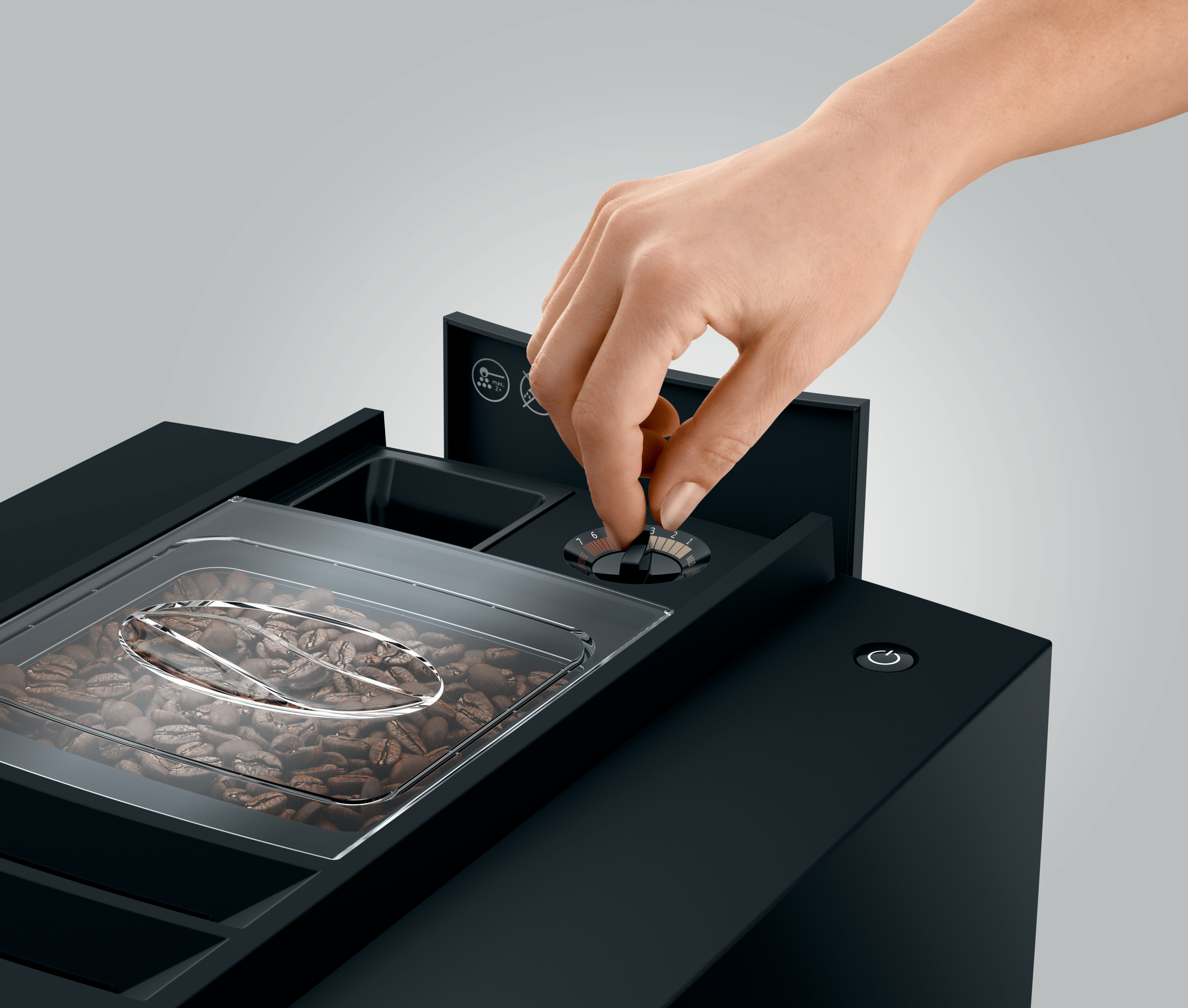 Eine Hand dreht einen Drehknopf an einer schwarzen Kaffeemaschine. Kaffeebohnen sind in einem transparenten Behälter sichtbar.