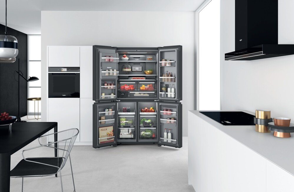 Otwarta lodówka z jedzeniem. Biała kuchnia z czarnym stołem i okapem.