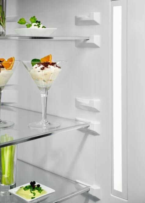 Wnętrze lodówki ze szklanymi półkami, deserami i pionowym światłem.