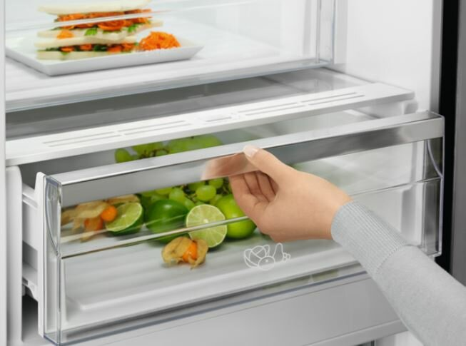Ręka otwiera szufladę w lodówce wypełnionej owocami i warzywami.