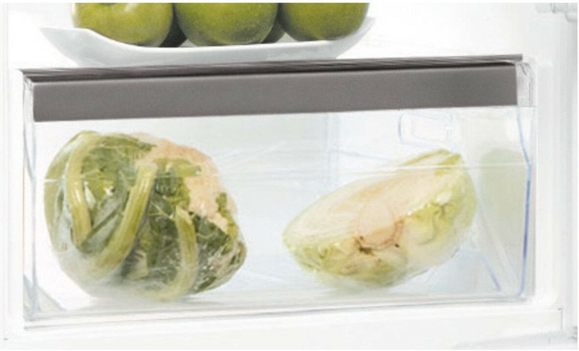 W szufladzie lodówki dwie zapakowane sałaty. Powyżej biała taca z zielonymi jabłkami.