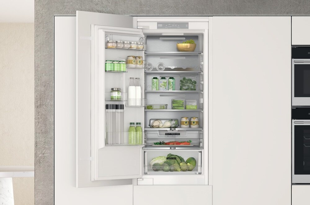 Biała lodówka z otwartymi drzwiami. Wewnątrz znajdują się półki z produktami spożywczymi, jak słoiki i owoce.