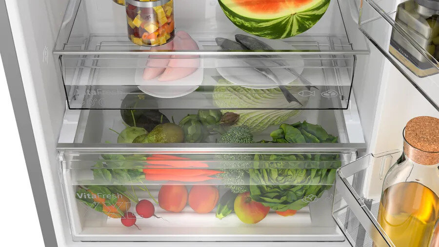 Wnętrze lodówki z szufladami. Widoczne owoce, warzywa, pikle i butelki z sokiem.