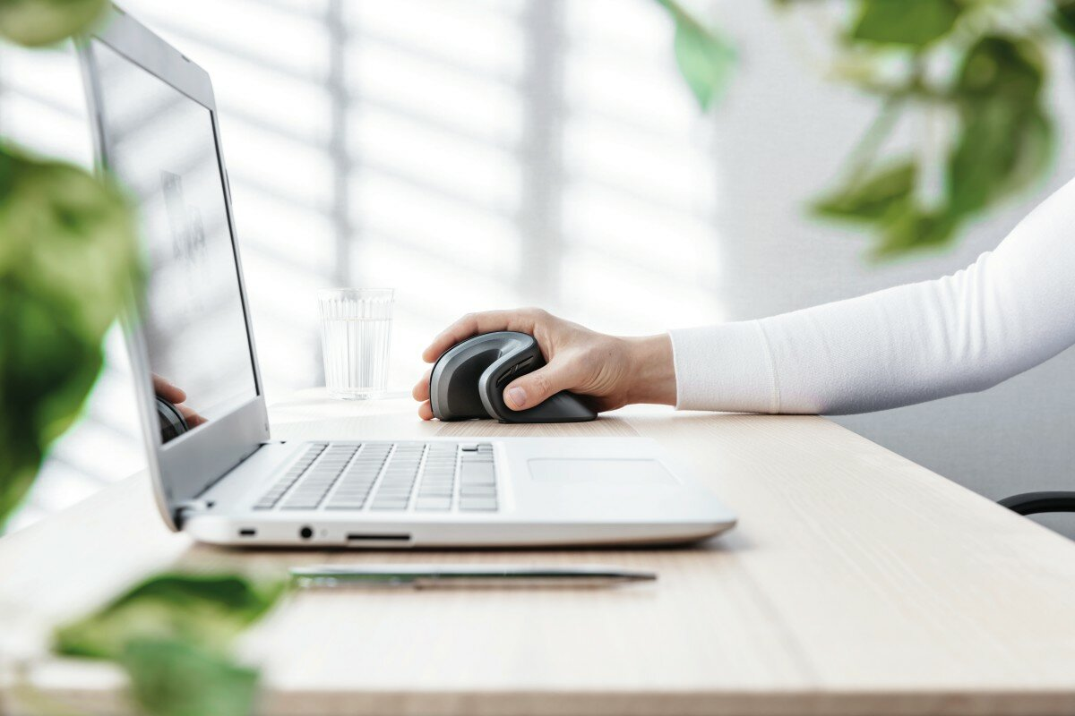 Dłoń osoby używającej pionowej, ergonomicznej myszy na drewnianym biurku.