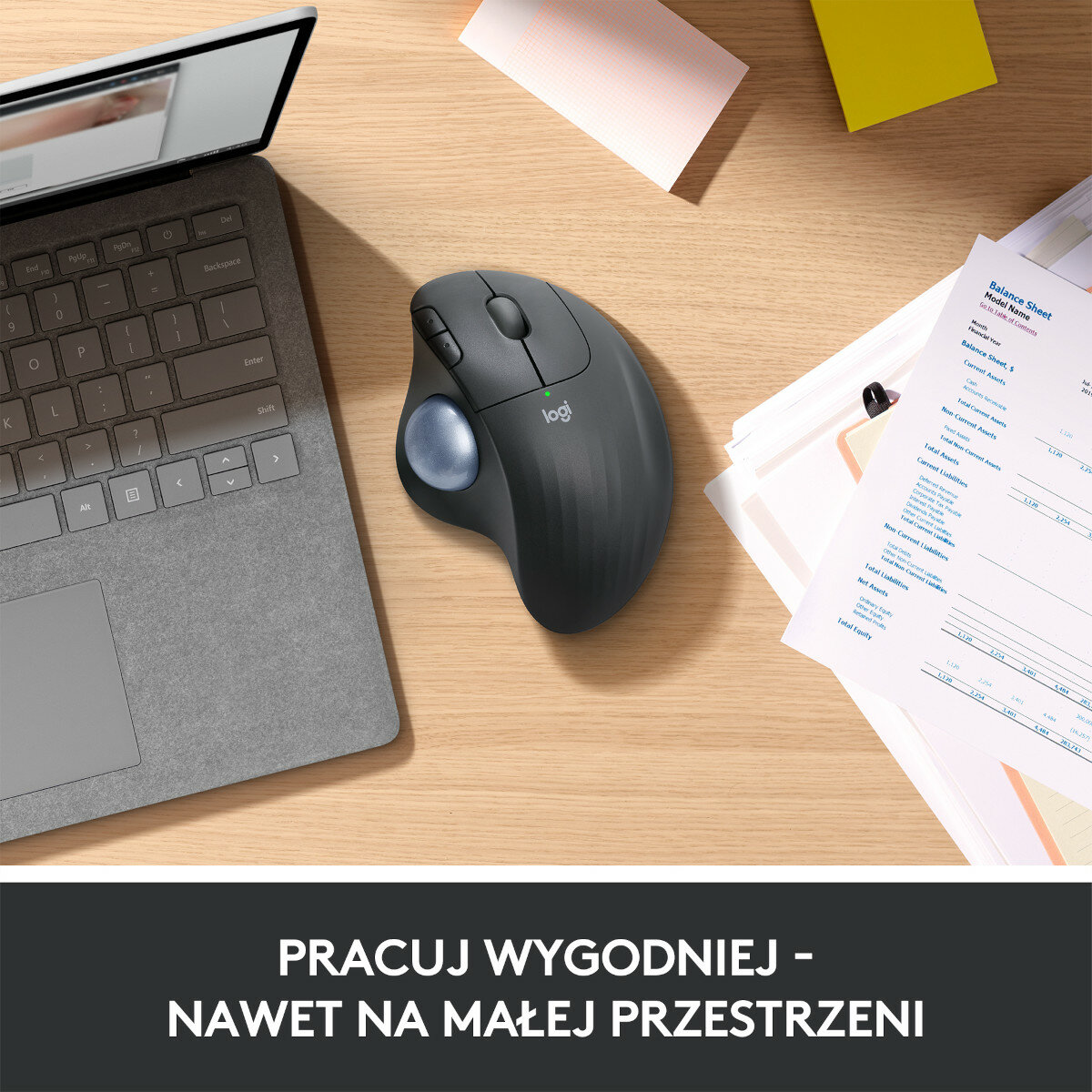 Czarna ergonomiczna mysz z niebieską kulką na drewnianym biurku, obok laptopa i dokumentów.