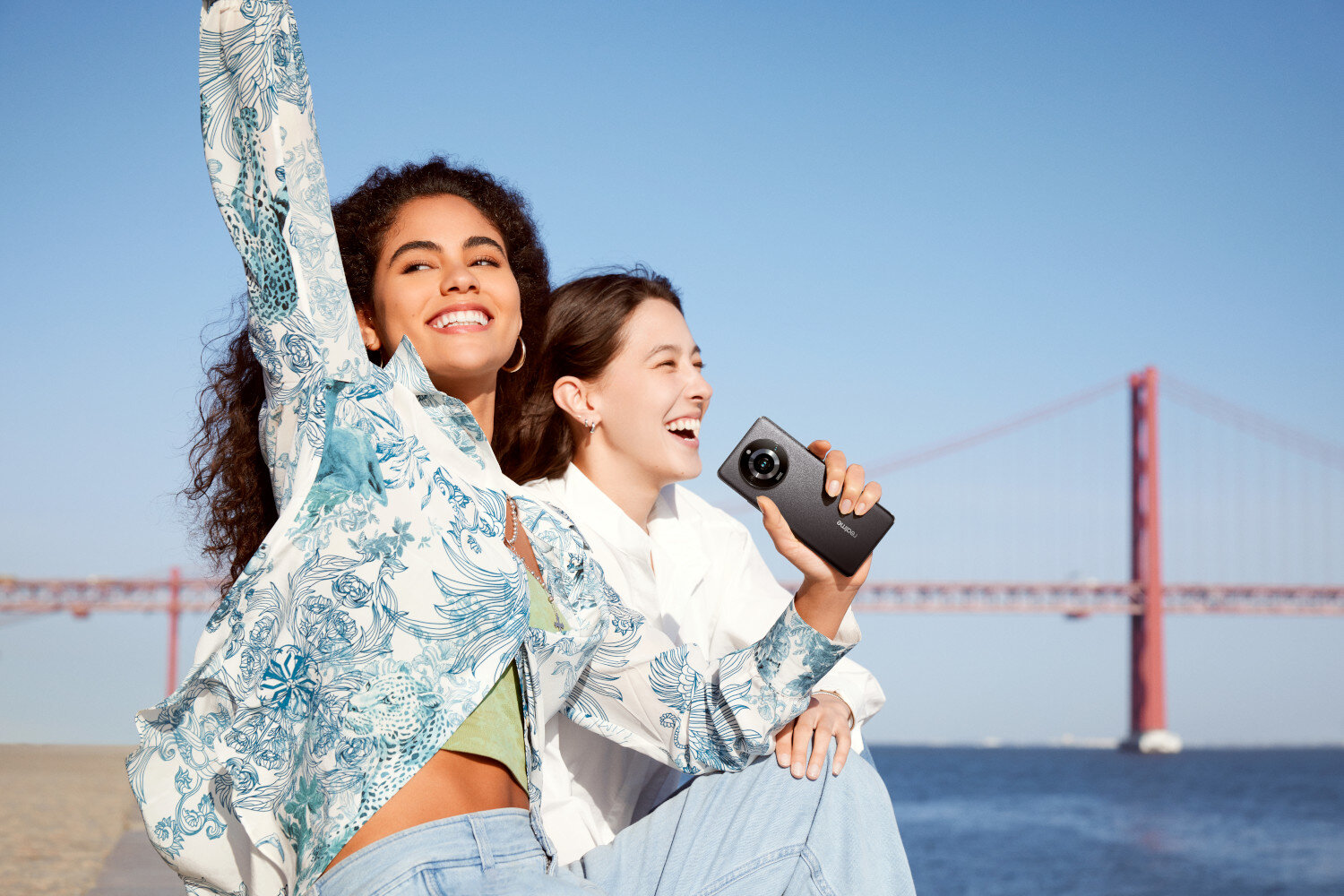 Dwie uśmiechnięte kobiety trzymające telefon, z mostem i niebieskim niebem w tle.