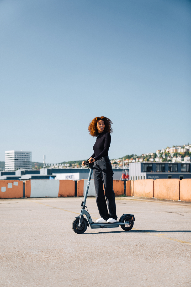 Donna in sella a uno scooter azzurro. Sorride di fronte agli edifici.
