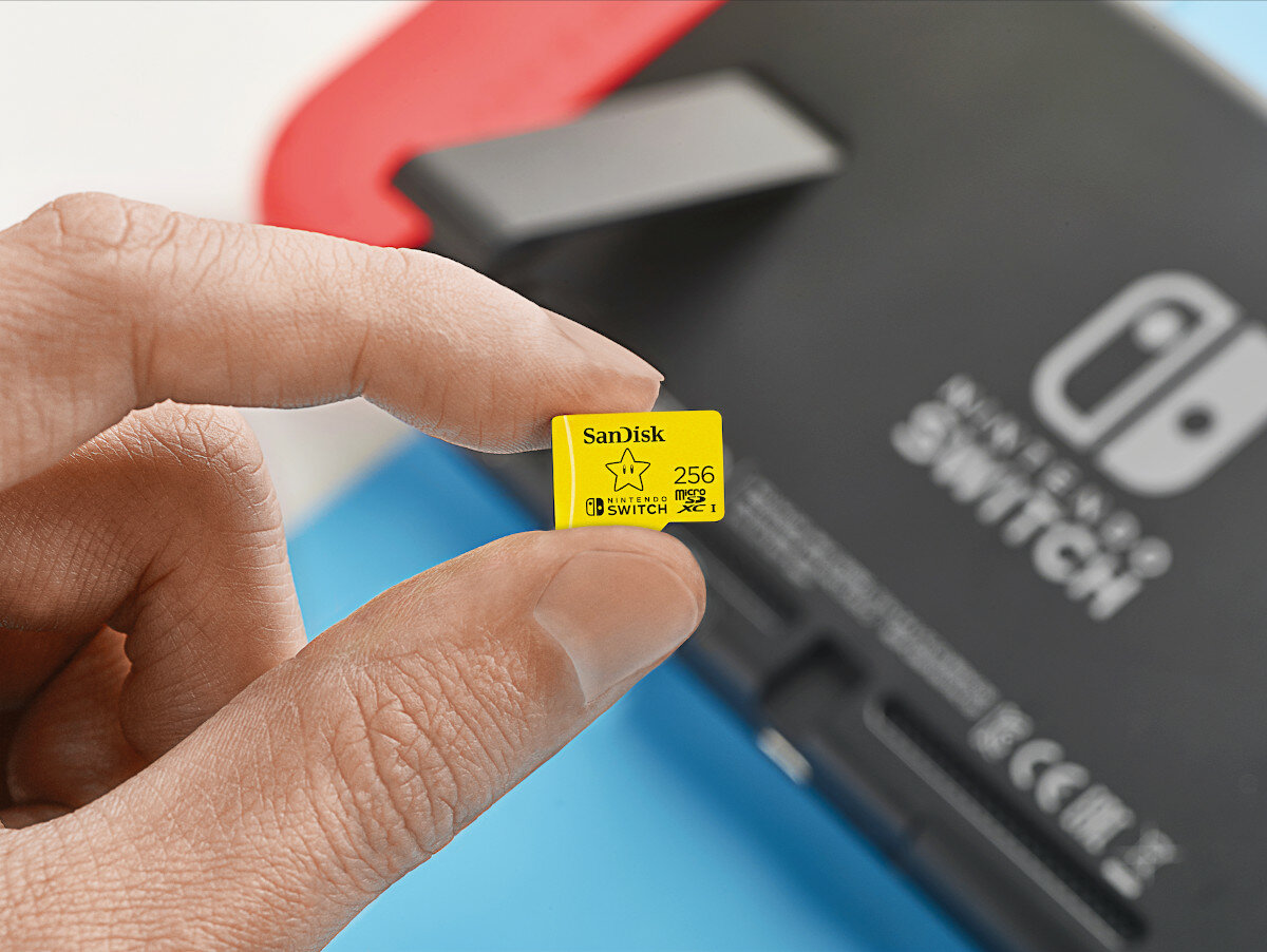 Ręka trzymająca żółtą kartę microSD SanDisk obok konsoli Nintendo Switch.