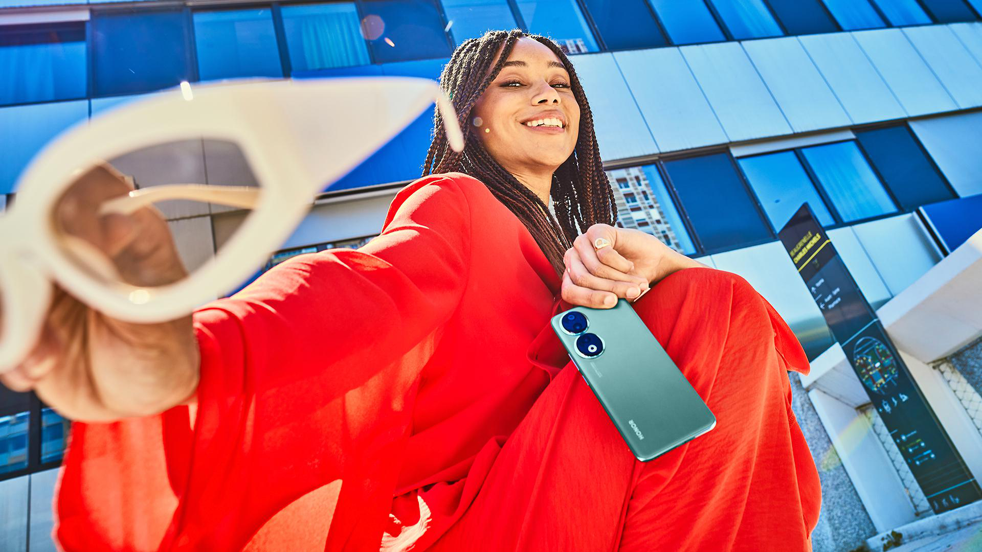 Eine Frau in Rot, die ein grünes Telefon und eine Sonnenbrille hält, mit Stadt im Hintergrund.