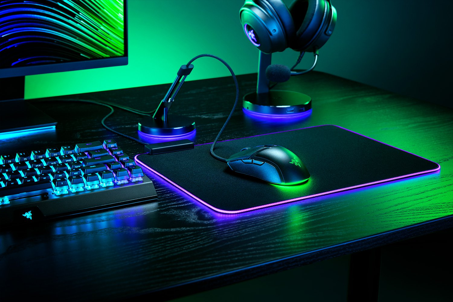 Gaming-Setup: Maus, Mauspad, Tastatur, Headset und Monitor auf einem schwarzen Holztisch mit grüner Beleuchtung.