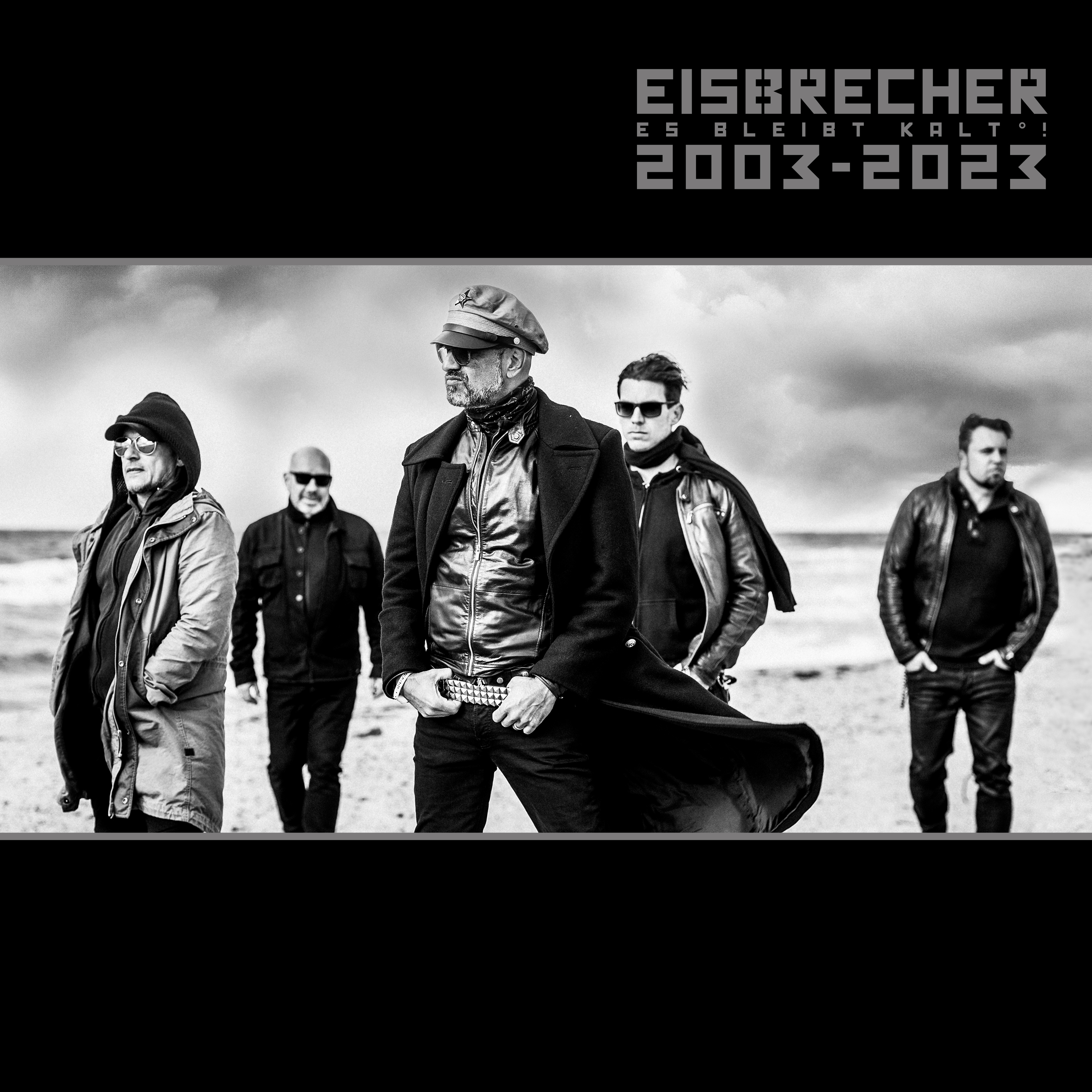 Band am Strand, Schwarz-Weiß-Foto. Männer in dunklen Mänteln und Hüten. Bewölkter Himmel im Hintergrund.