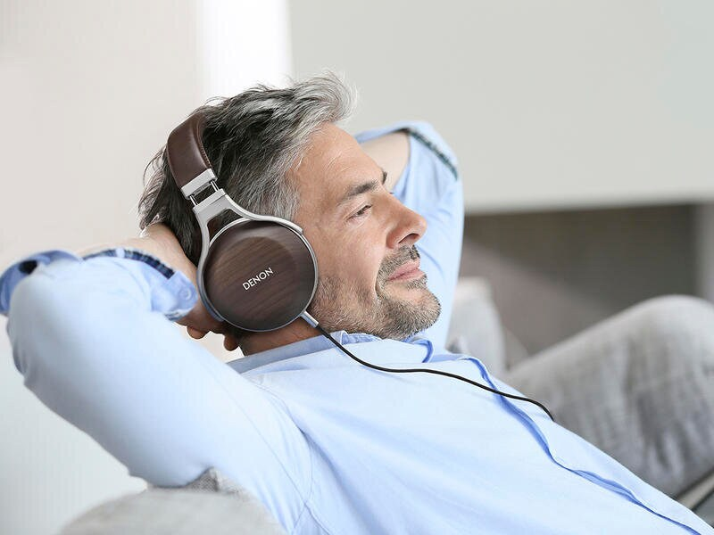 Mężczyzna w niebieskiej koszuli w brązowych słuchawkach Denon, zrelaksowany z rękami za głową. Jasne tło.