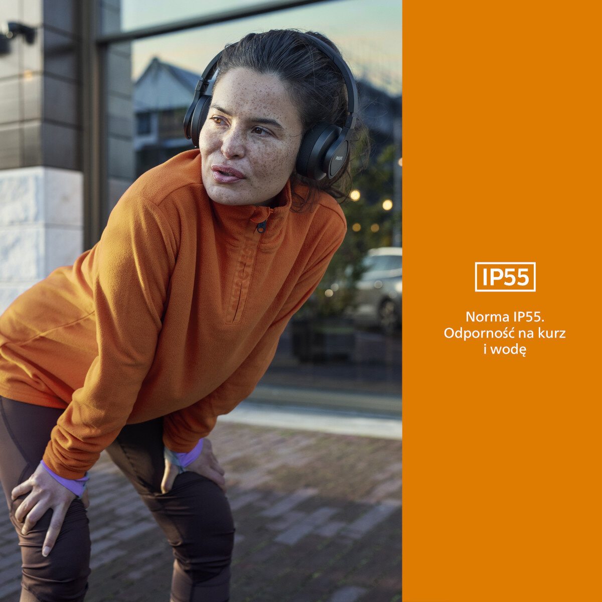 Kobieta w pomarańczowej bluzie z słuchawkami, stojąca na zewnątrz.