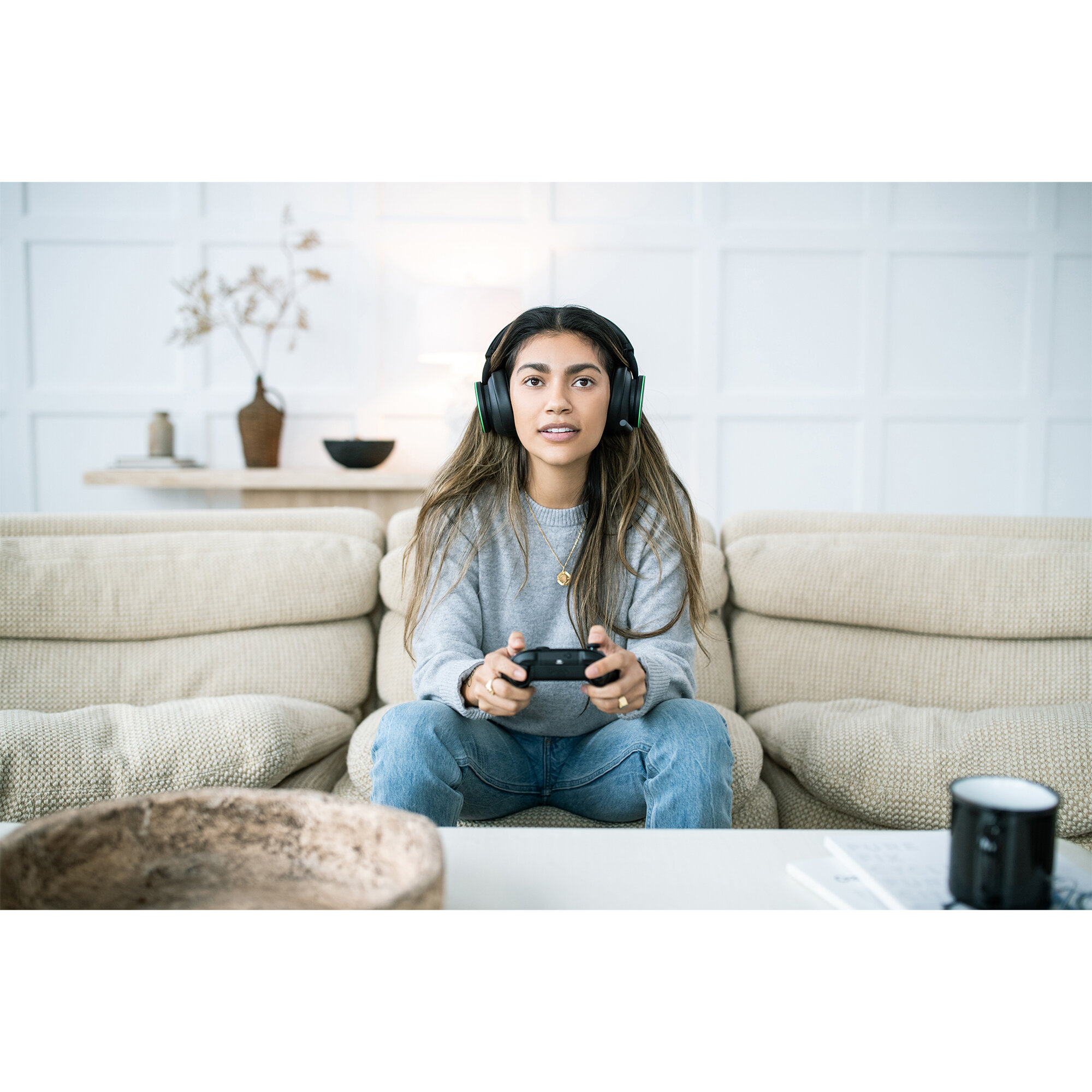 Młoda kobieta ze słuchawkami i kontrolerem siedzi na kanapie, patrząc w kamerę. Przed nią znajduje się stolik kawowy.