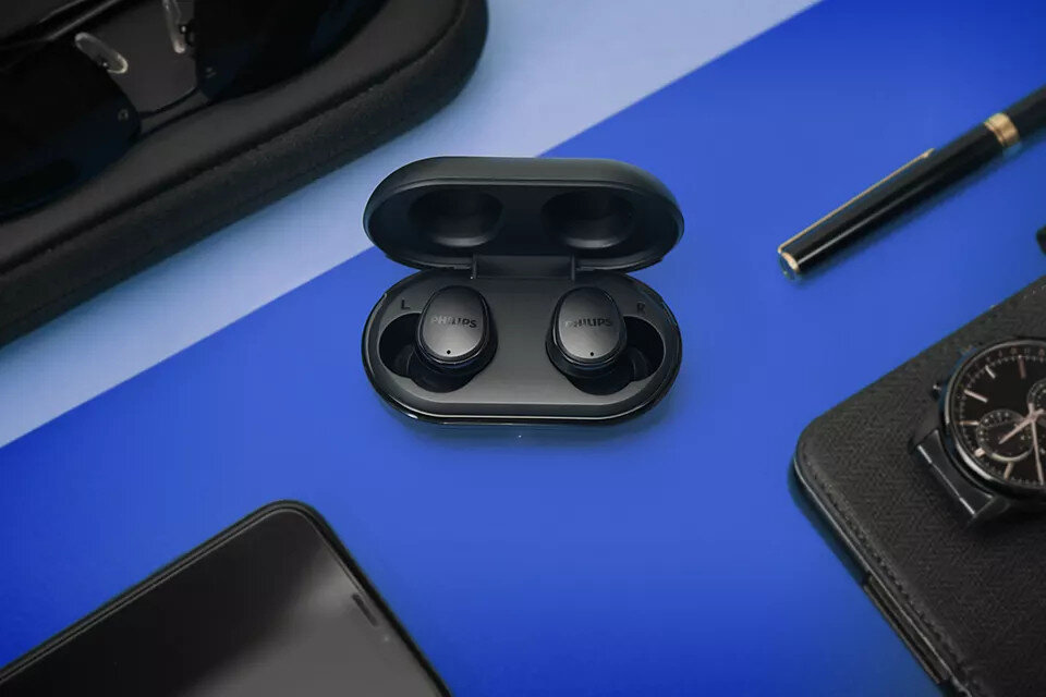 Czarne słuchawki douszne w etui ładującym, z okularami, długopisem i zegarkiem na niebieskiej powierzchni.