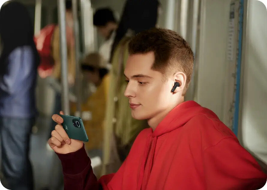 Mężczyzna w czerwonej bluzie z kapturem używa słuchawek, trzymając zielony smartfon w metrze.