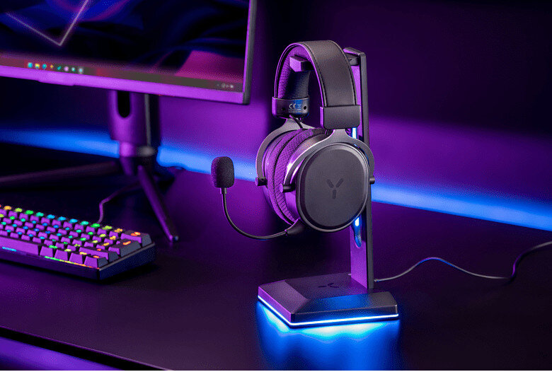 Zestaw słuchawkowy do gier na stojaku, z klawiaturą i monitorem, na ciemnej powierzchni z fioletowym oświetleniem.