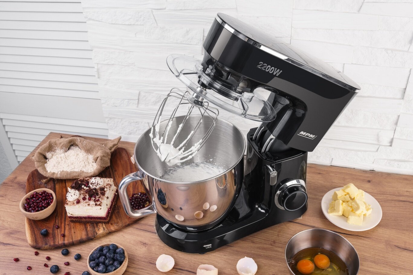 Czarny mikser planetarny z trzepaczką ubijającą śmietanę, obok składników takich jak mąka, ciasto, jagody i masło.