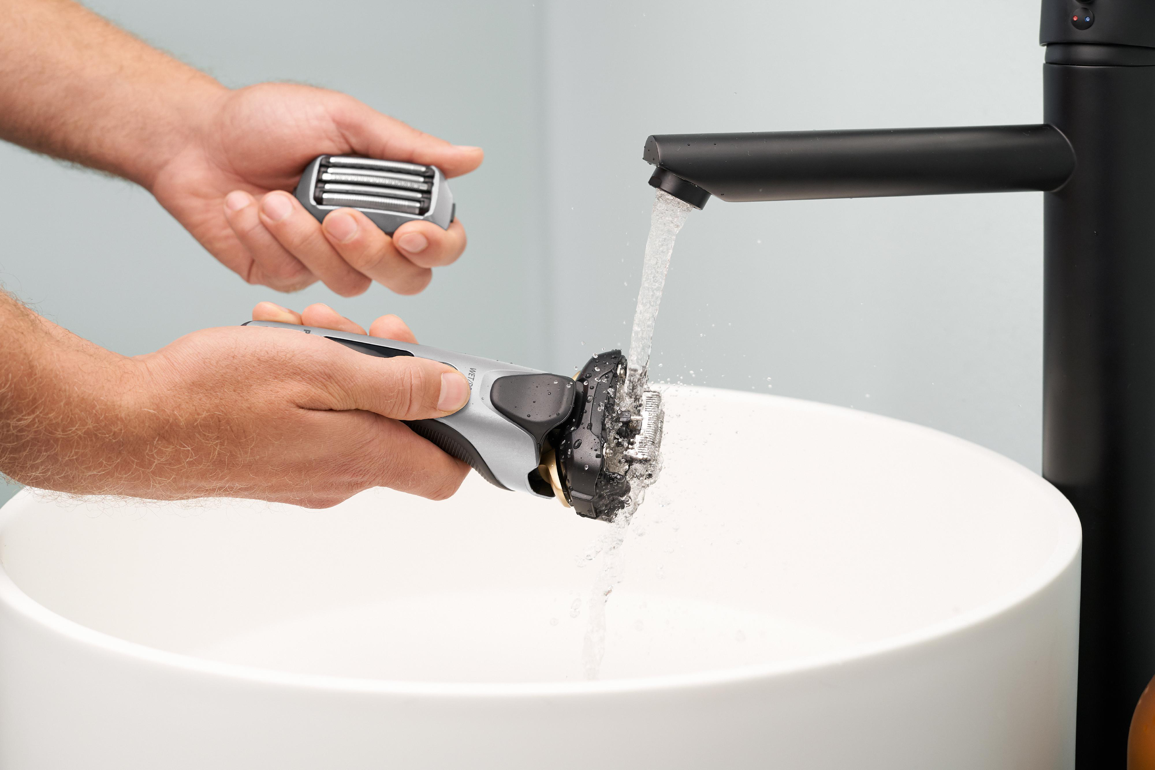 Hände waschen einen elektrischen Rasierer. Wasser fließt aus einem schwarzen Wasserhahn in ein Waschbecken.