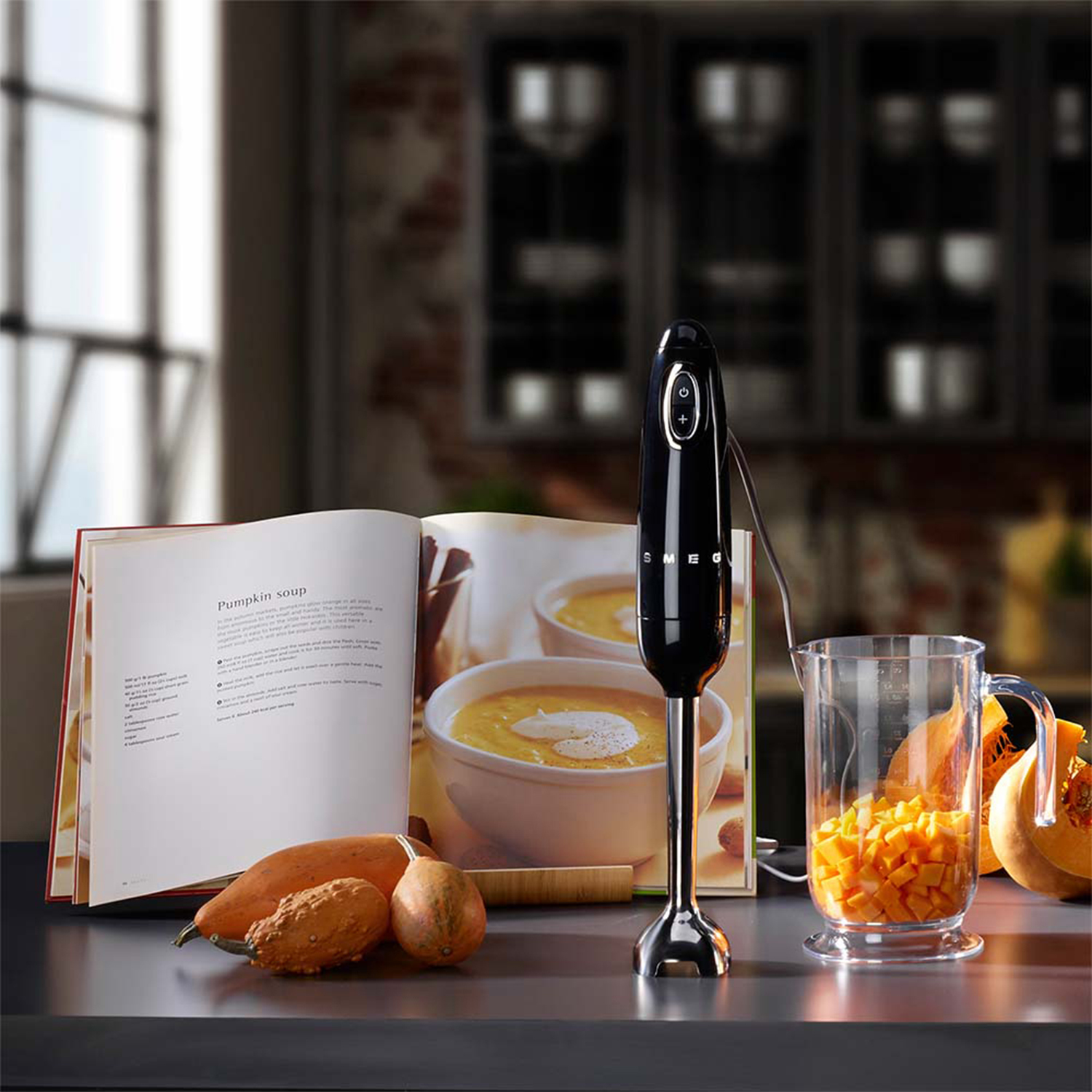 Un frullatore a immersione nero accanto a un libro di cucina aperto, zucche e zucca tritata in un barattolo.