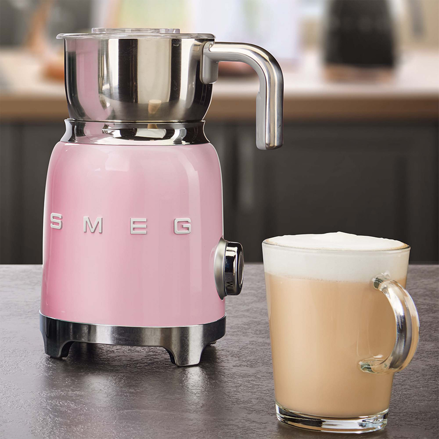 Un montalatte SMEG rosa e un bicchiere di cappuccino con manico sono su un tavolo.