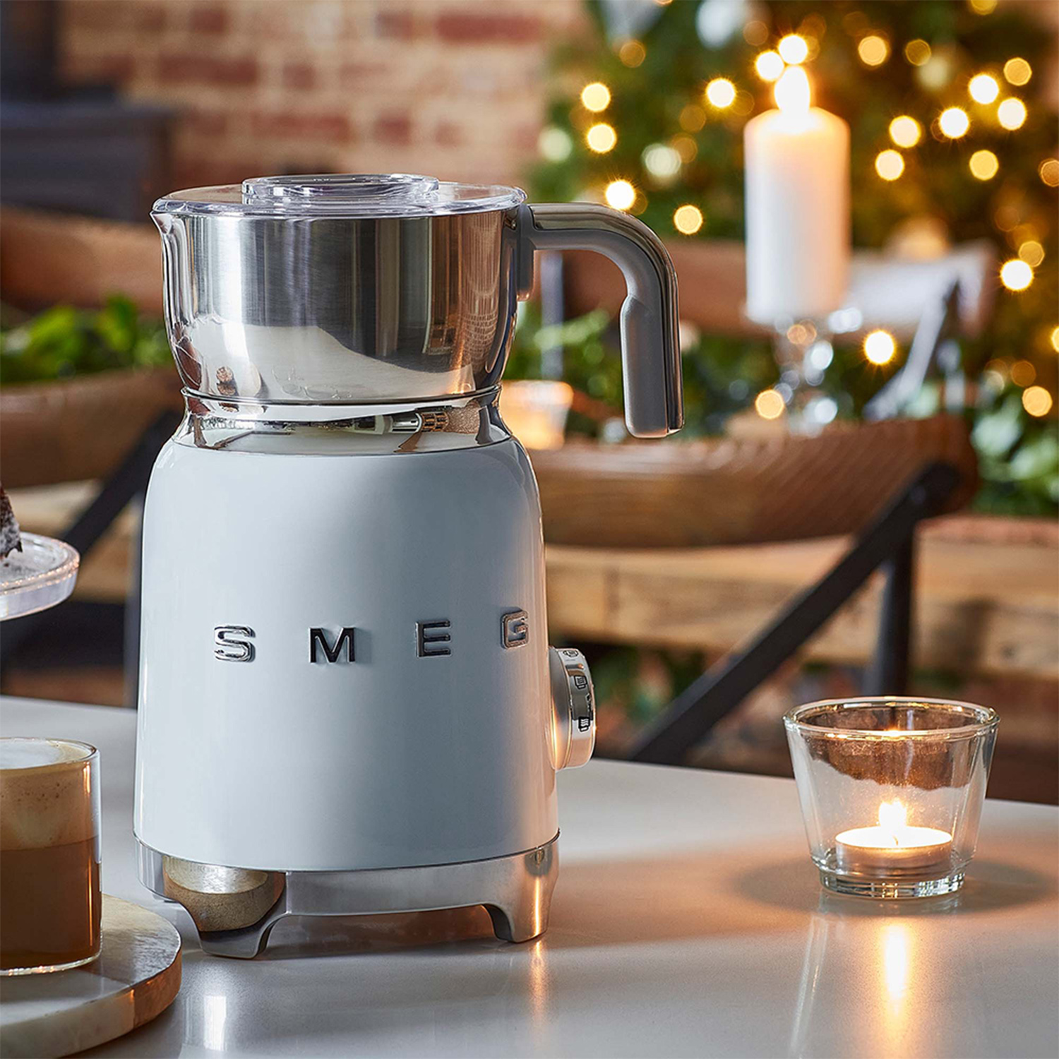 Un montalatte SMEG bianco e argento su un tavolo. Una candela accesa e un bicchiere in primo piano.