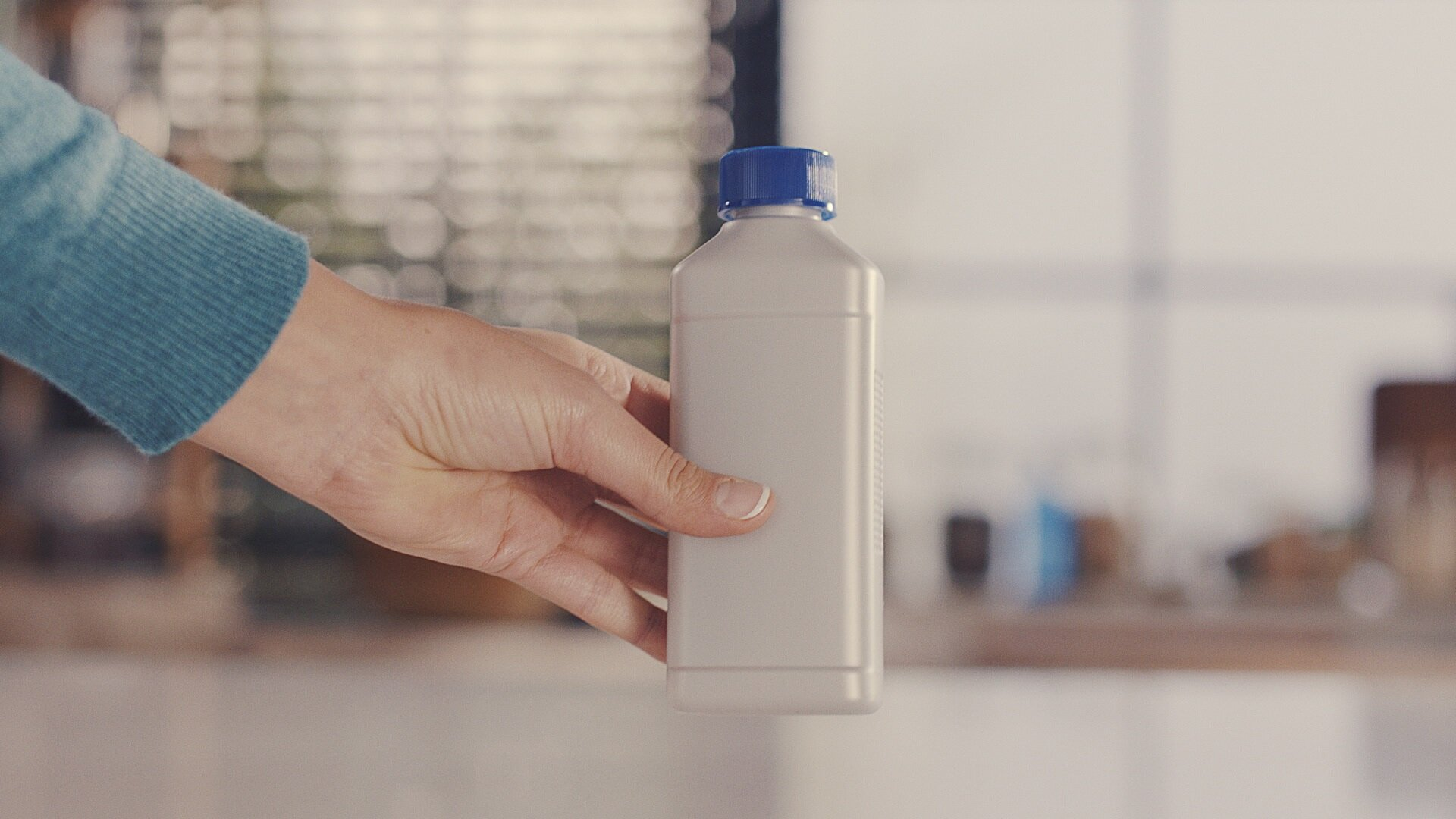 Ręka trzymająca białą butelkę z niebieską zakrętką. Rozmyte tło.
