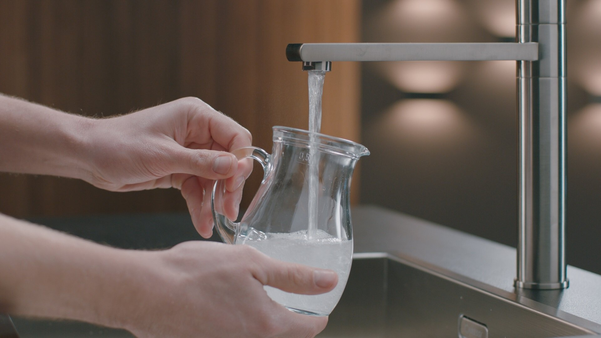 Dłonie napełniają przezroczysty szklany dzbanek wodą z kranu nad zlewem ze stali nierdzewnej.