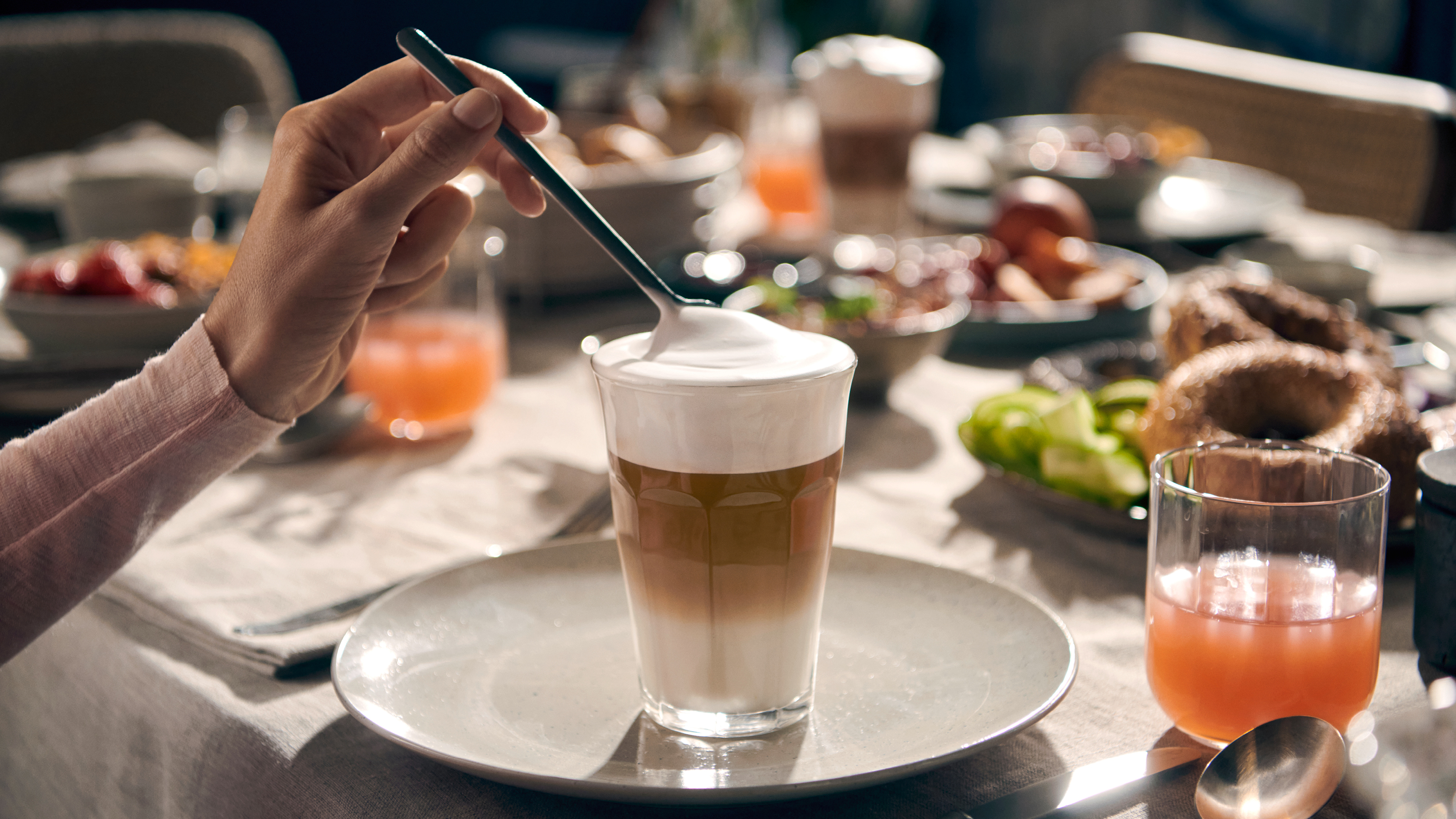 Ein Latte wird zubereitet. Eine Hand rührt den Schaum auf dem Latte in einem Glas um.