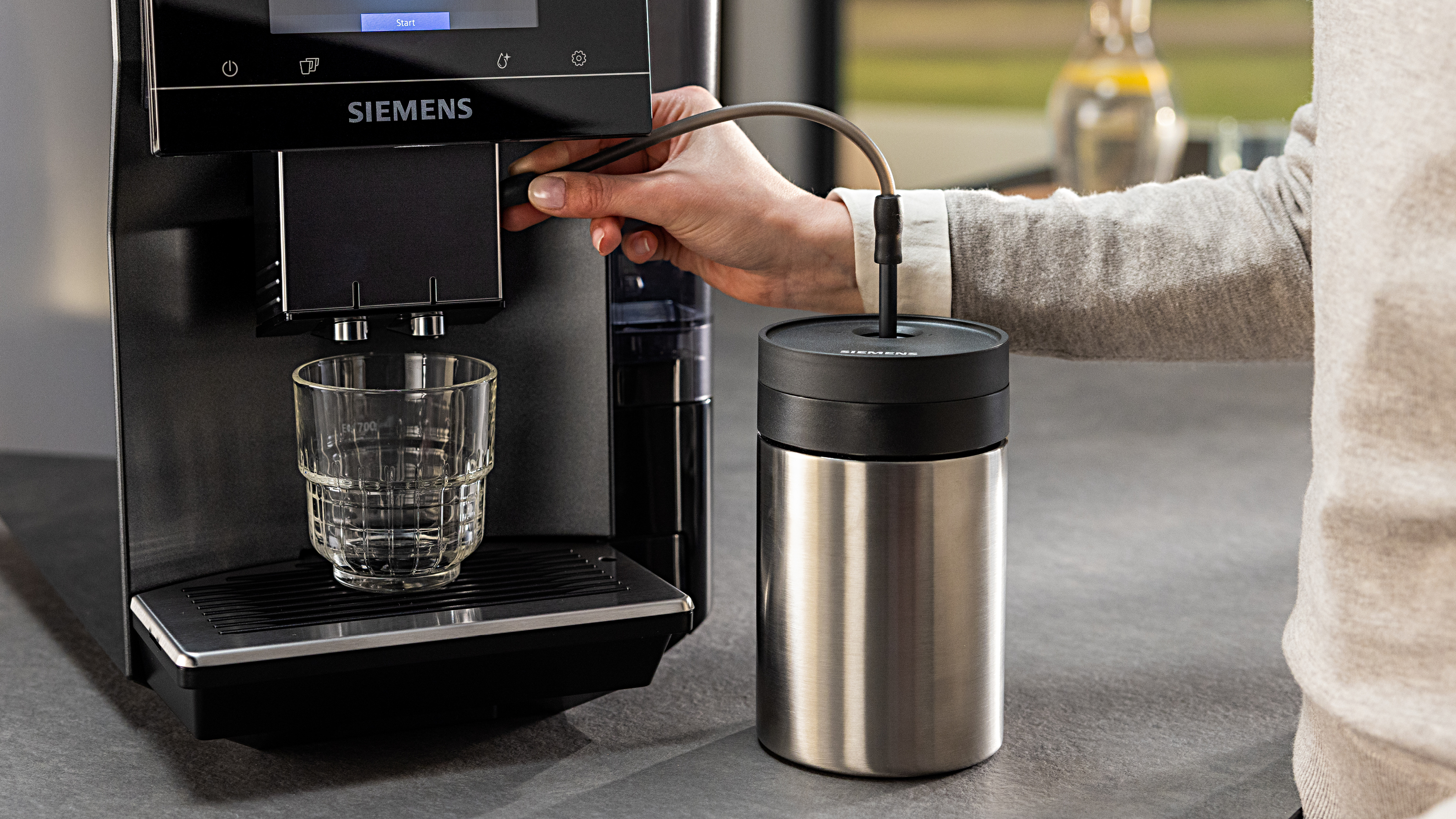 Eine Person benutzt eine Kaffeemaschine, gießt Kaffee in ein Glas, mit Milchaufschäumer.