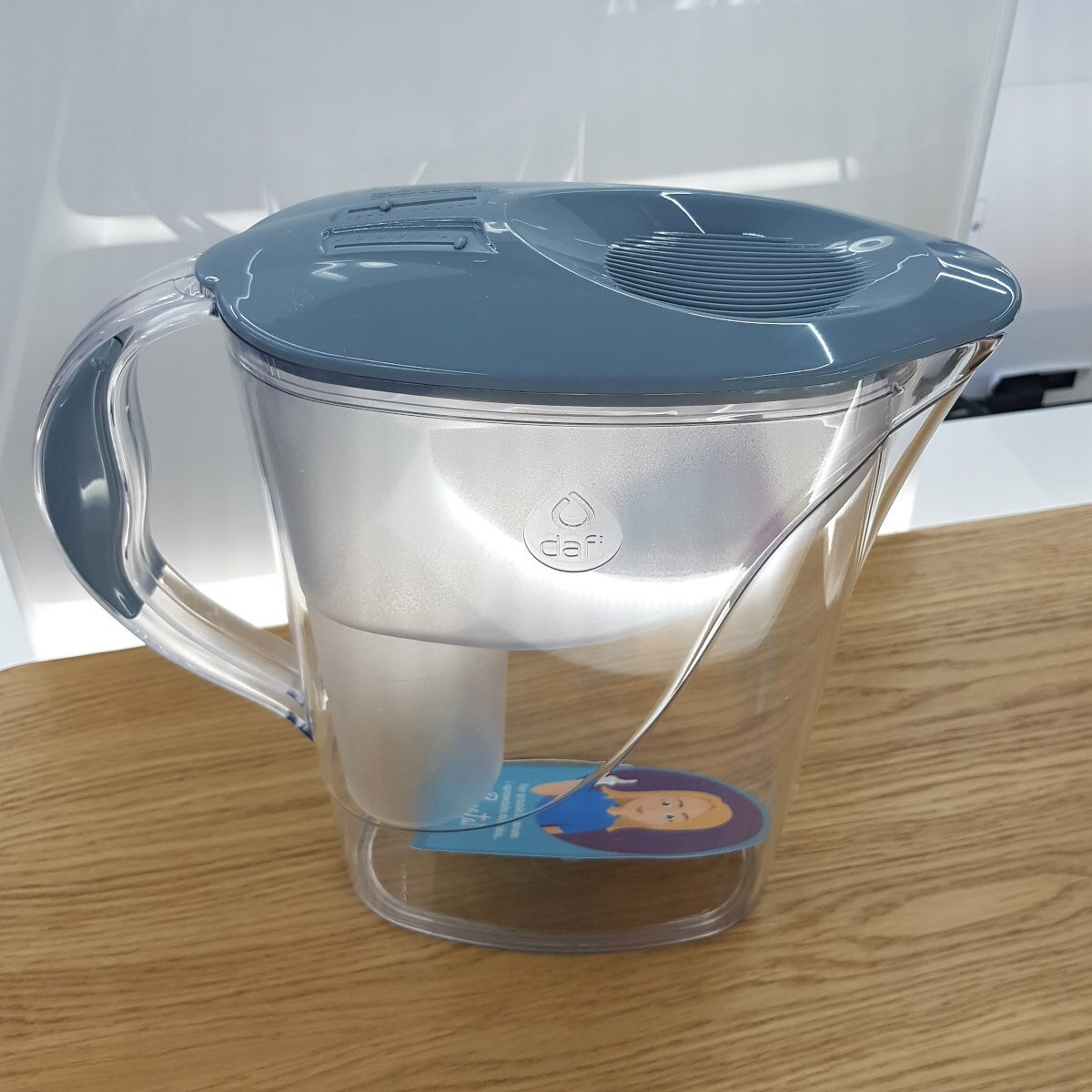 Przezroczysty dzbanek z filtrem do wody z szarą pokrywką, napełniony wodą, na drewnianej powierzchni.