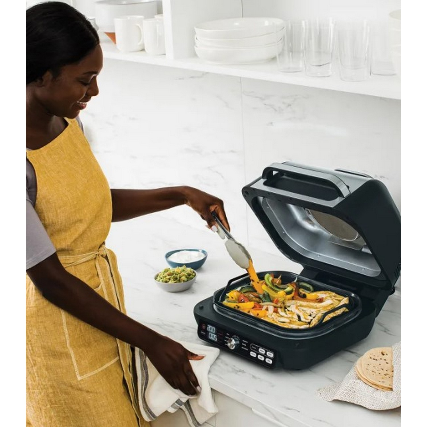 A woman with a yellow apron is using tongs to move food on a grill.