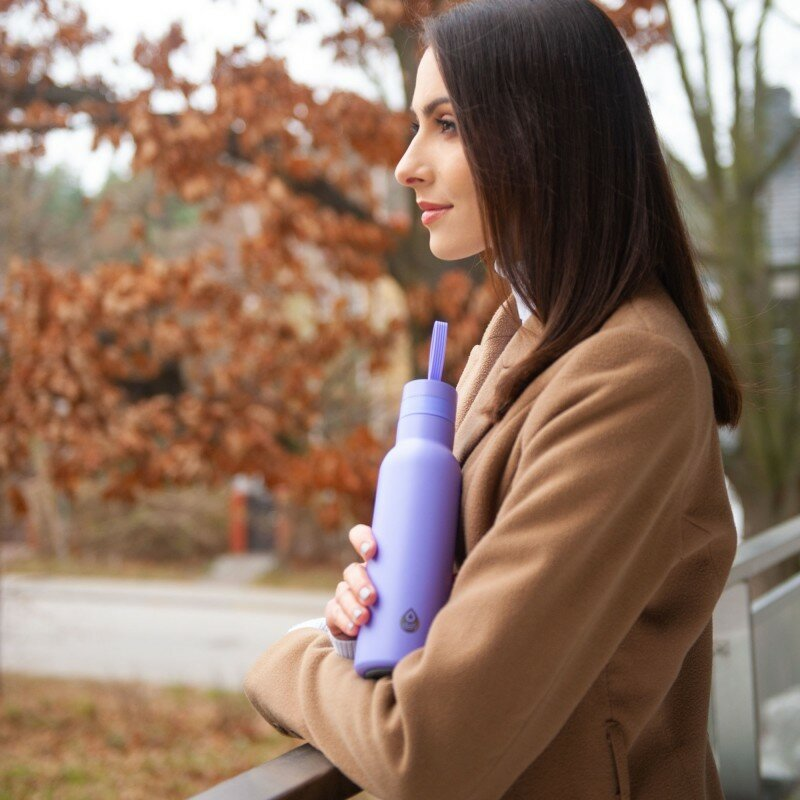 Kobieta z ciemnymi włosami trzyma na zewnątrz fioletową butelkę wody ze słomką.
