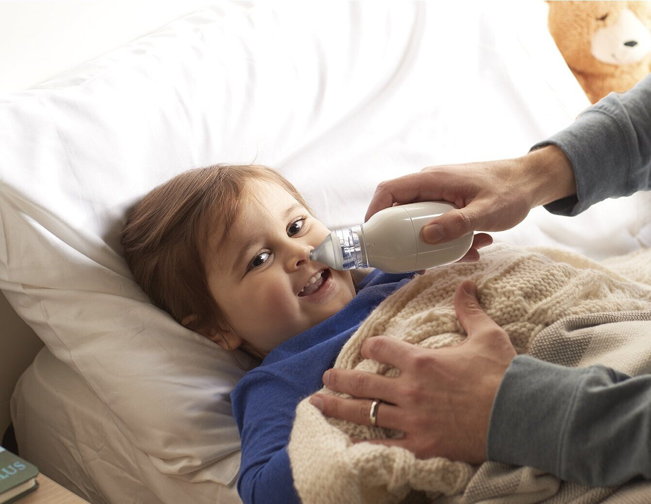 Dziecko leżące w łóżku, leczone aspiratorem do nosa przez osobę dorosłą.