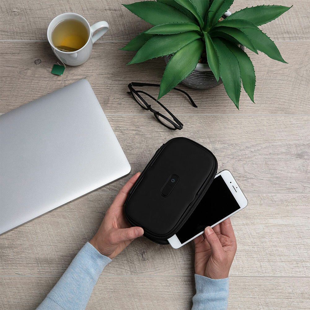 Ręce wkładają smartfon do czarnego etui obok laptopa, herbaty i okularów.