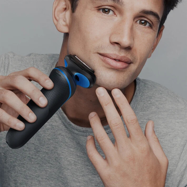 Mężczyzna goli twarz golarką elektryczną. Golarka jest czarno-niebieska.