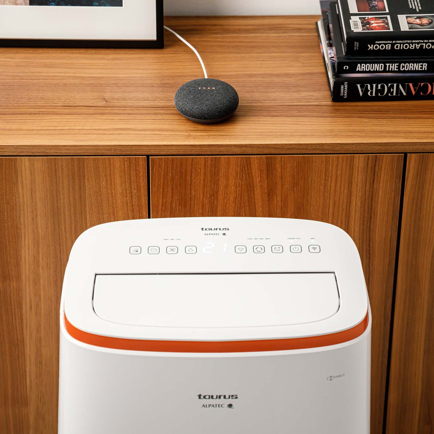 Un altavoz inteligente está sobre un mueble de madera, con libros y un purificador de aire en el fondo.