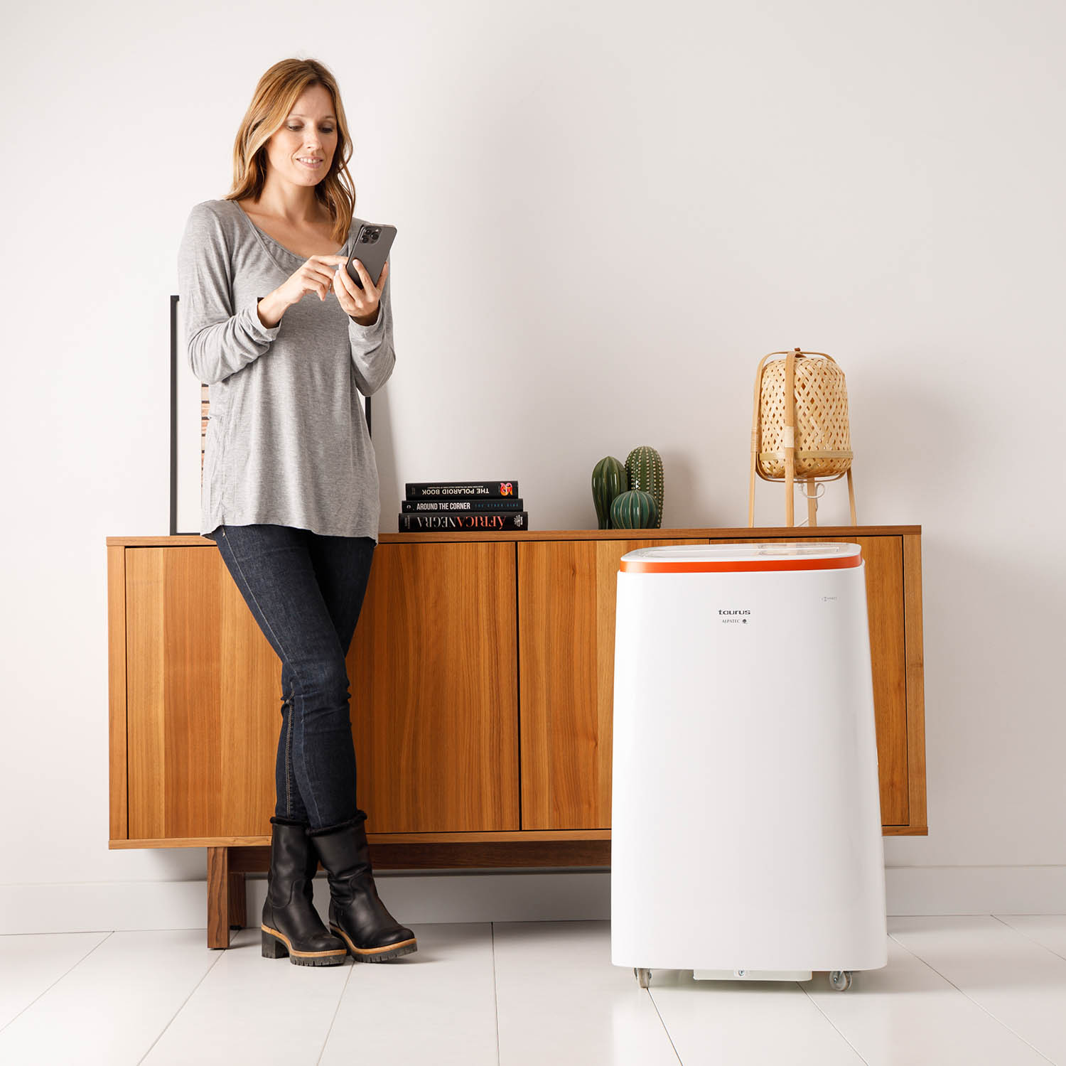 Mujer con teléfono junto a un armario de madera, purificador de aire al fondo. Cactus, lámpara visible.