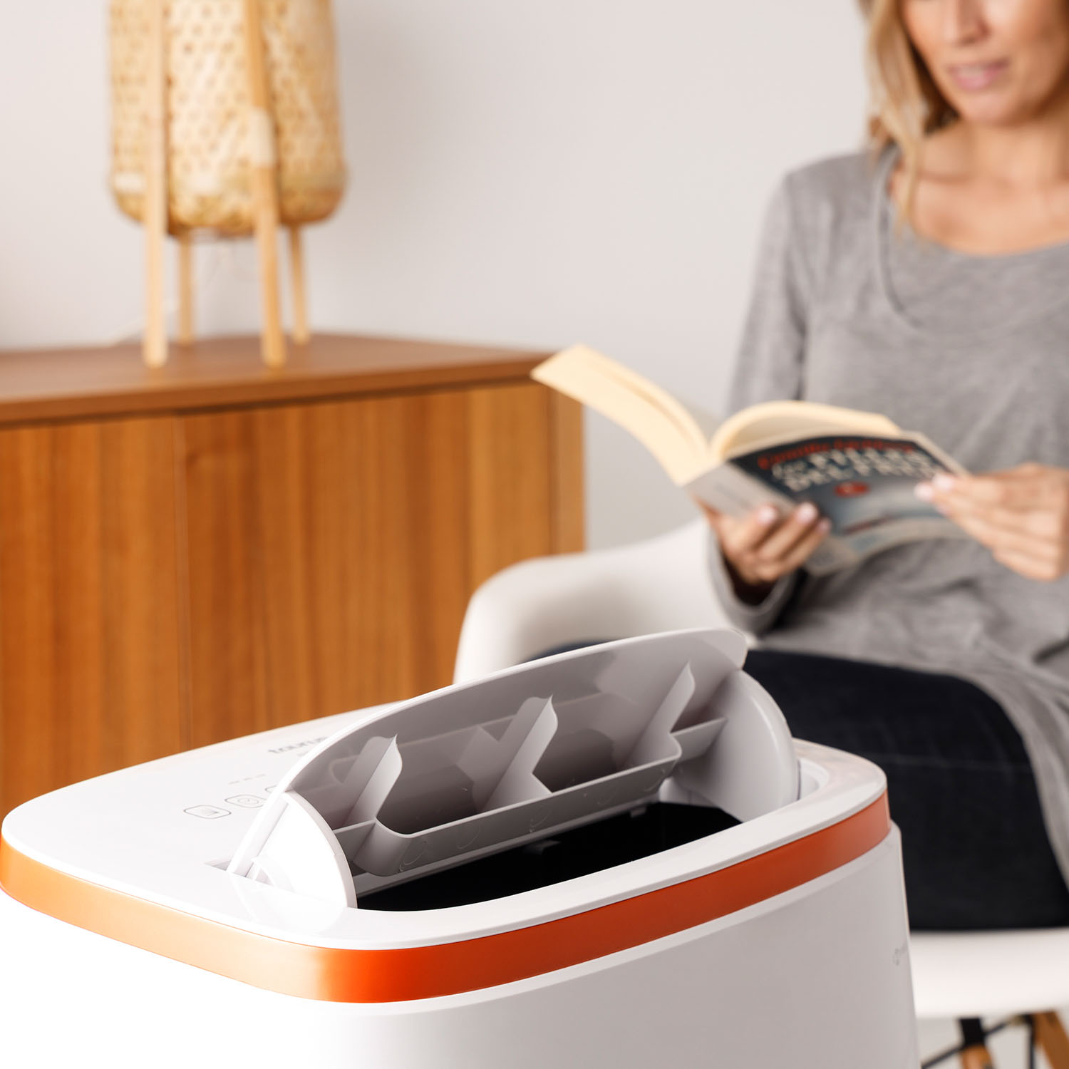 Mujer leyendo un libro cerca de un purificador de aire blanco y naranja. Abierto.