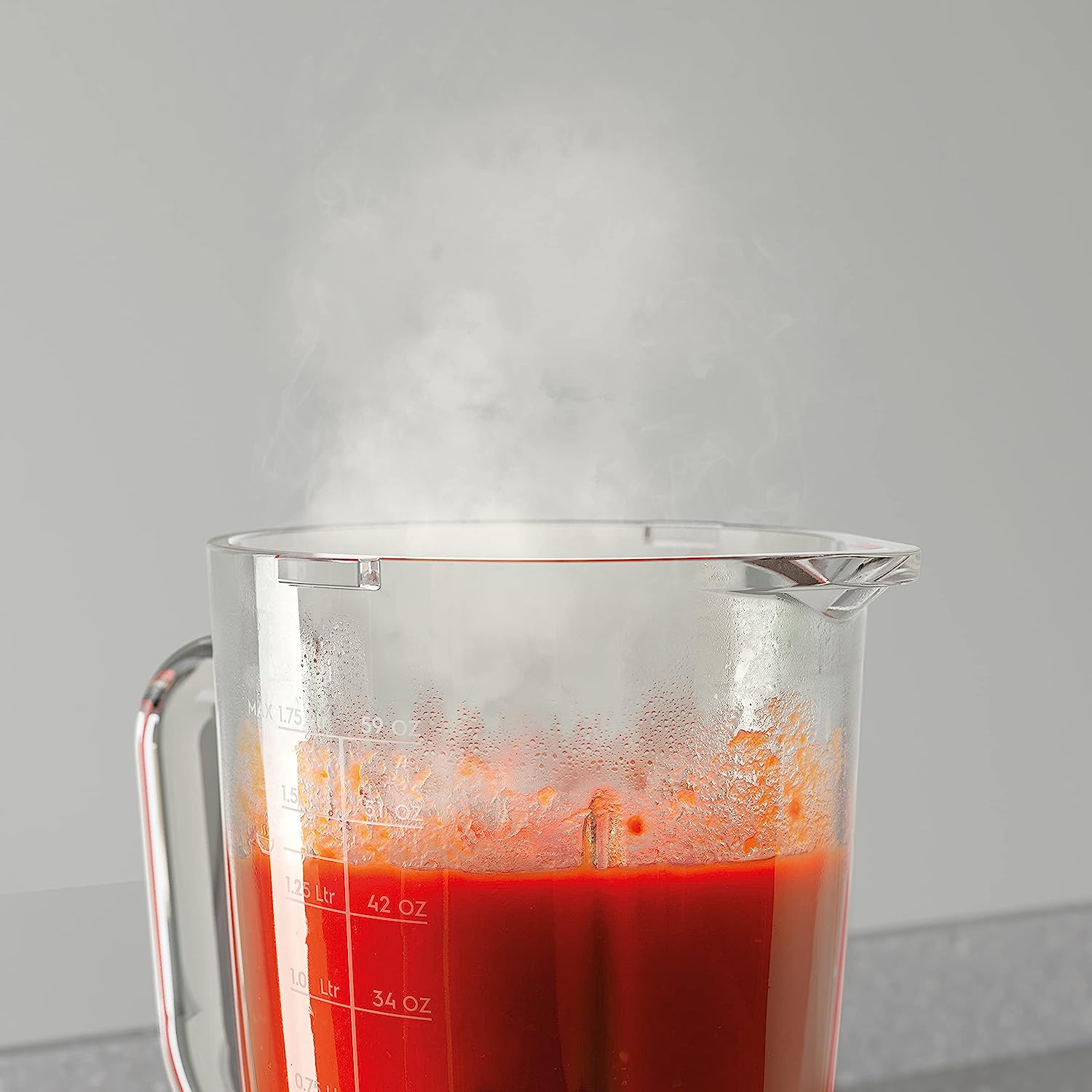 Kırmızı sıvı dolu şeffaf bir blender sürahisi. Buhar yükseliyor. Gri arka plan.