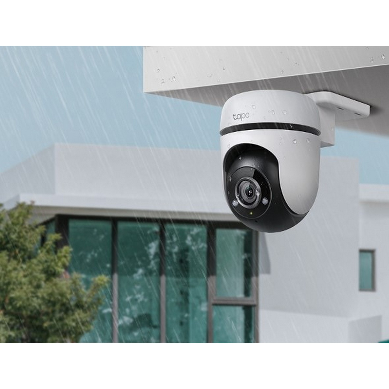 White and black security camera mounted under a white overhang, with rain falling and a house in background.