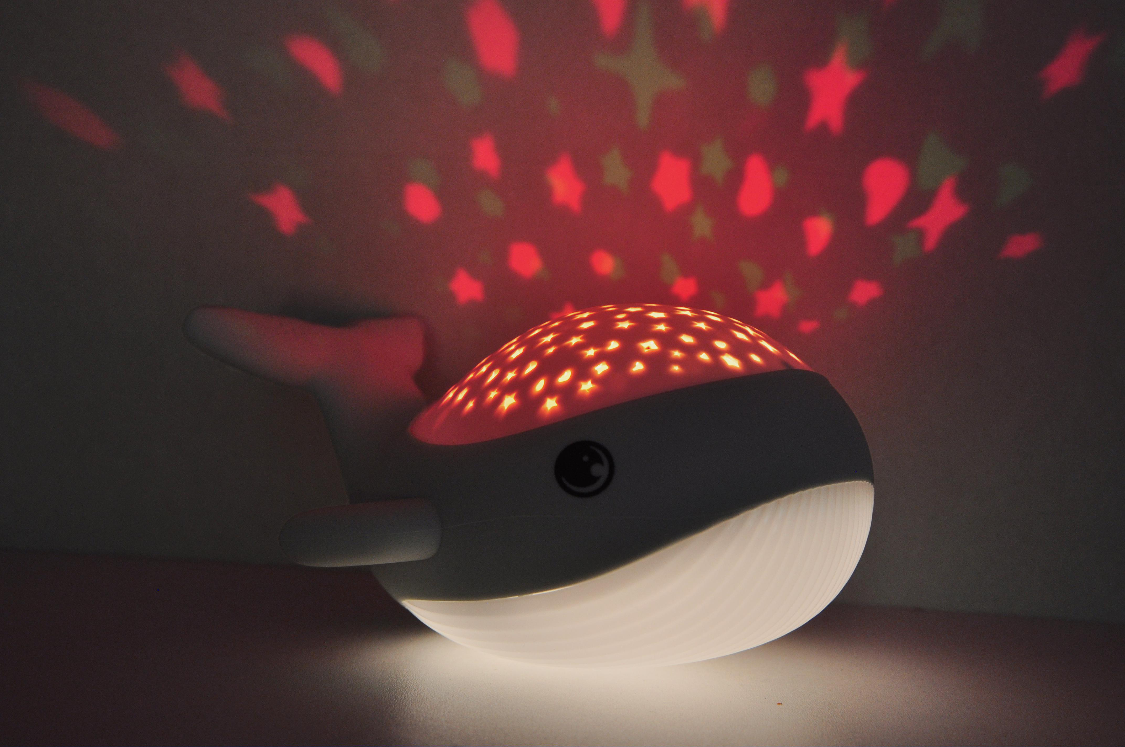Ein walförmiges Nachtlicht projiziert Sterne. Rote Sterne leuchten an der Wand. Das Licht ist weiß.